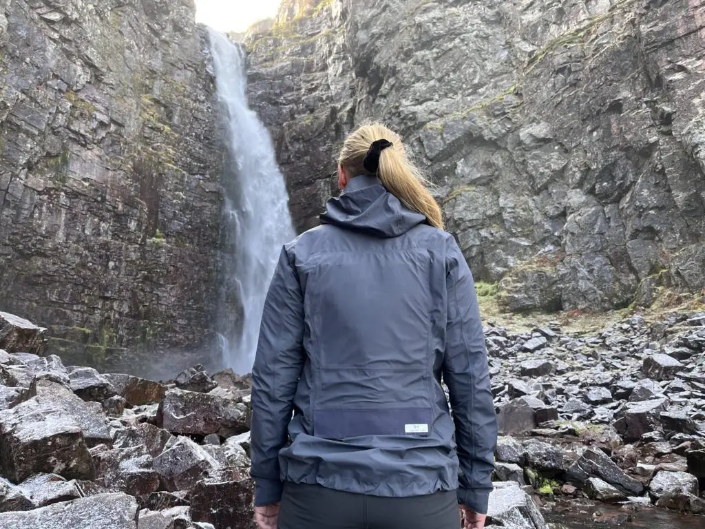 Photograph of a jacket, upgraded with LunaMicro's pump installed, being worn by a woman standing in front of a waterfall.